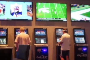 A row of self-service sports betting kiosks labeled 'Place Your Bets Here' inside a venue called 'The Gallery,' with two men using the machines while sports games are broadcast on television screens mounted on the wall above.