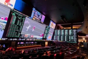 A modern, dimly lit casino sportsbook featuring a large, multi-screen video wall displaying live sports broadcasts and detailed betting odds for an American football game, with tiered theater-style seating for spectators.